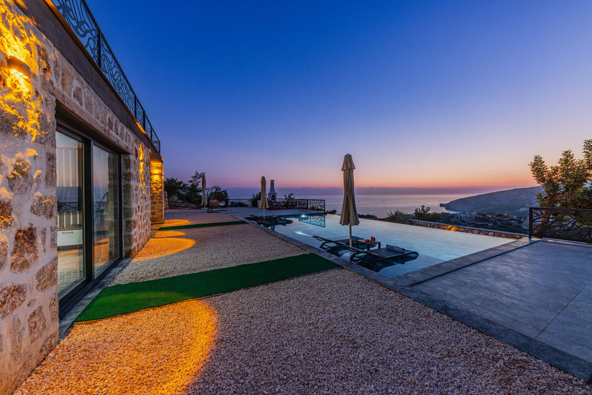 Terrasse extérieure en pierre avec piscine, éclairage chaud et vue dégagée vers la mer au coucher du soleil.