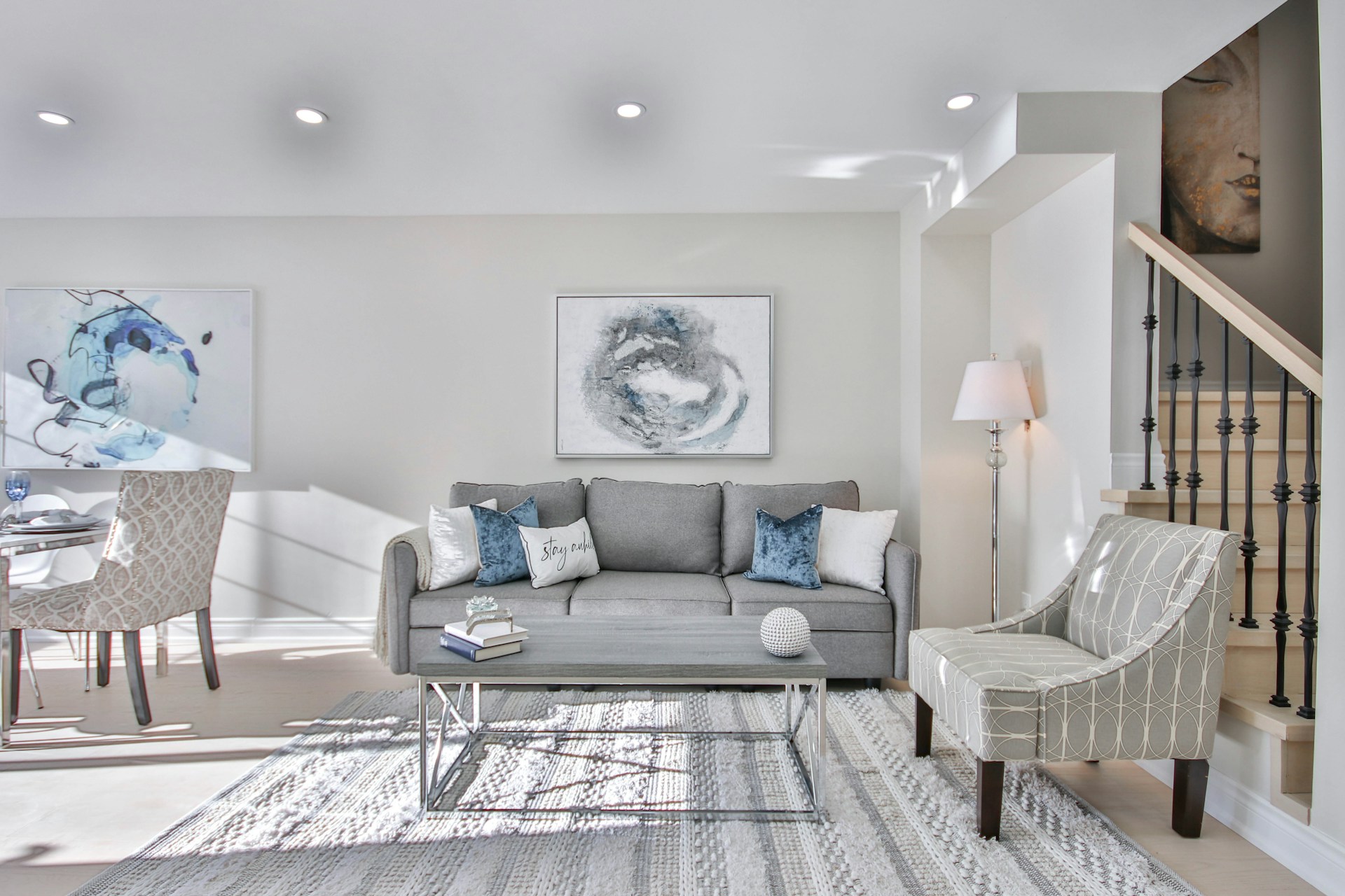 Salon gris baigné de lumière naturelle avec fauteuil à motif, tableaux abstraits et escalier latéral.