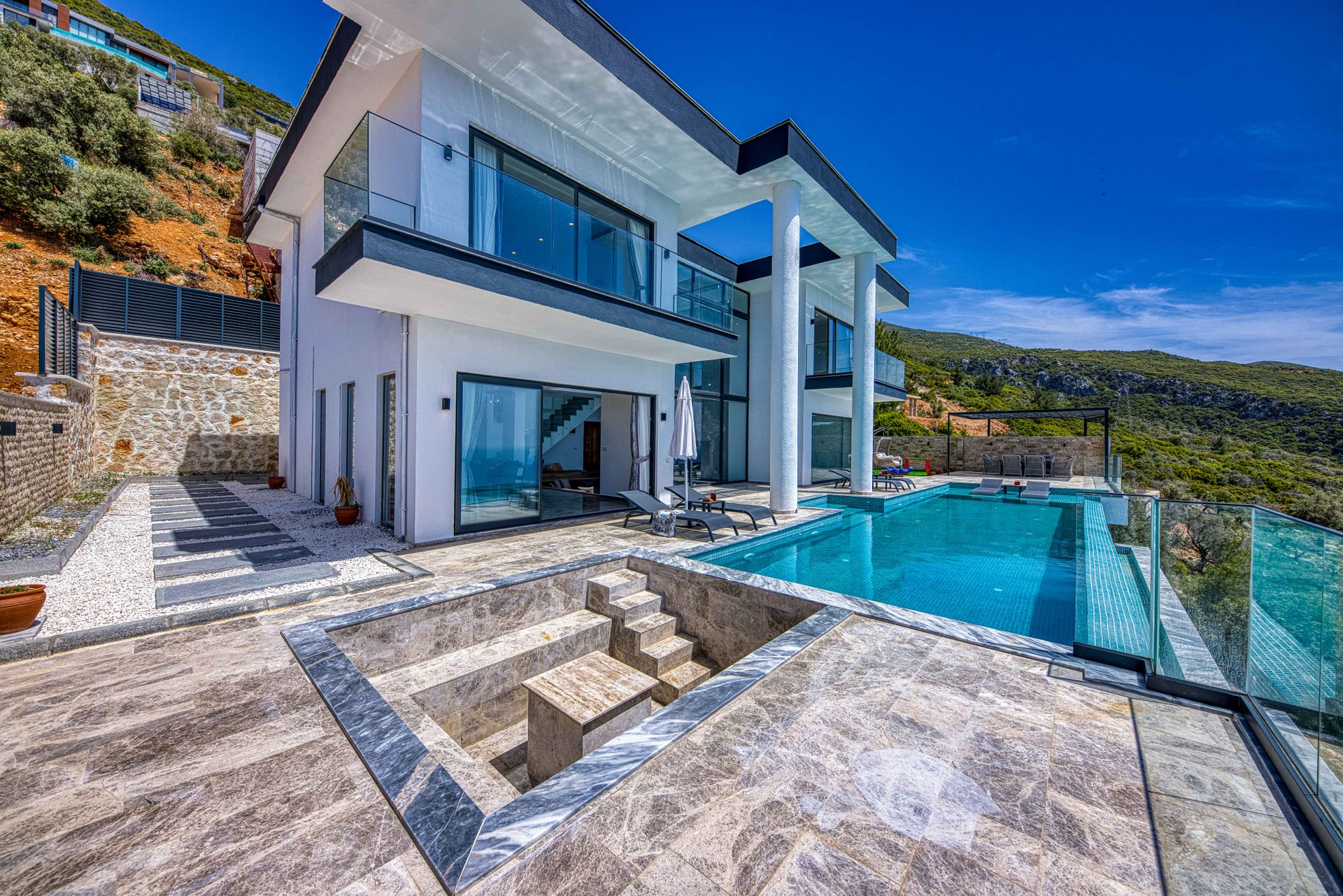 Façade contemporaine blanche avec terrasse minérale, piscine et collines en arrière-plan.