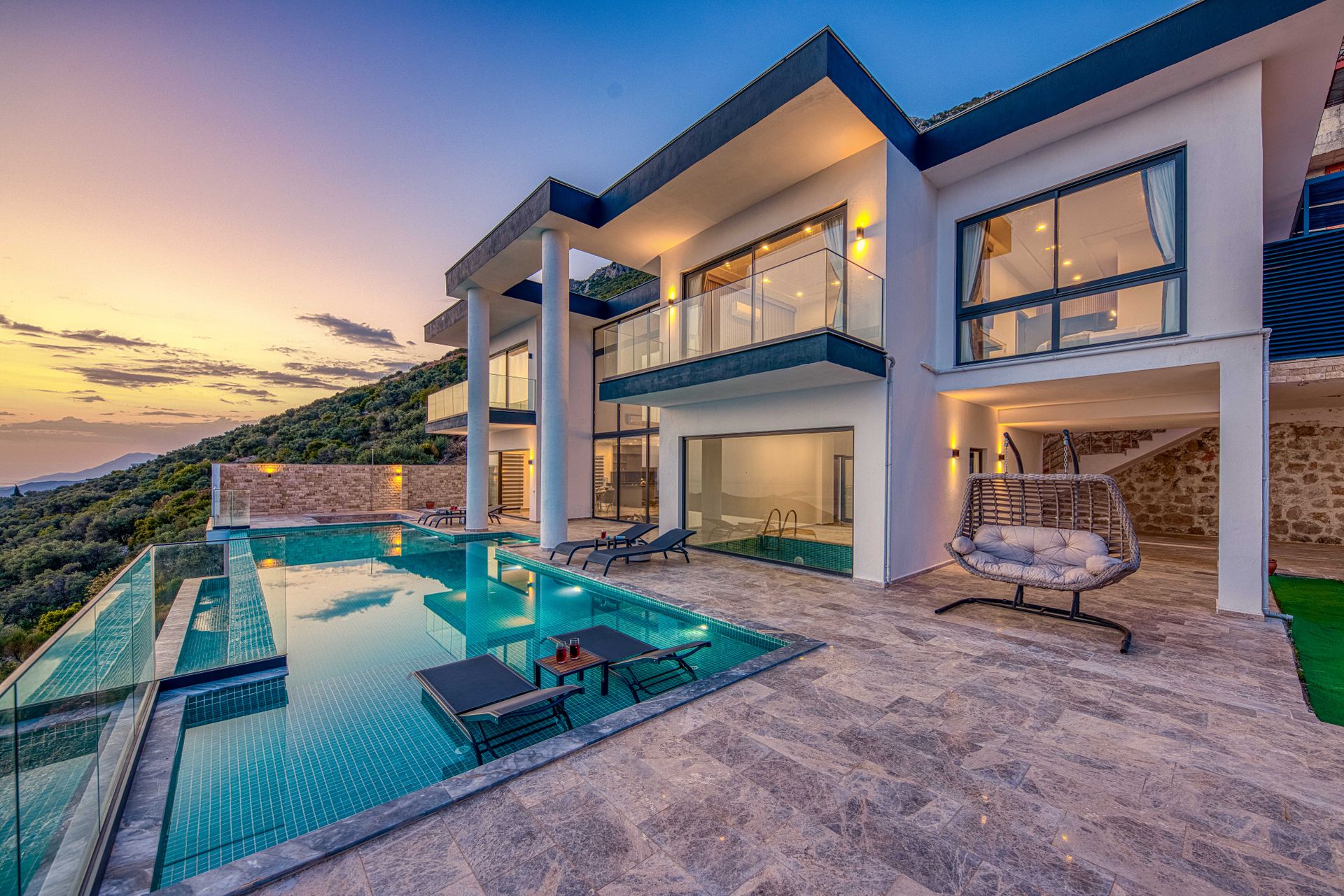 Villa moderne avec piscine au coucher du soleil, terrasse en pierre et fauteuil suspendu.